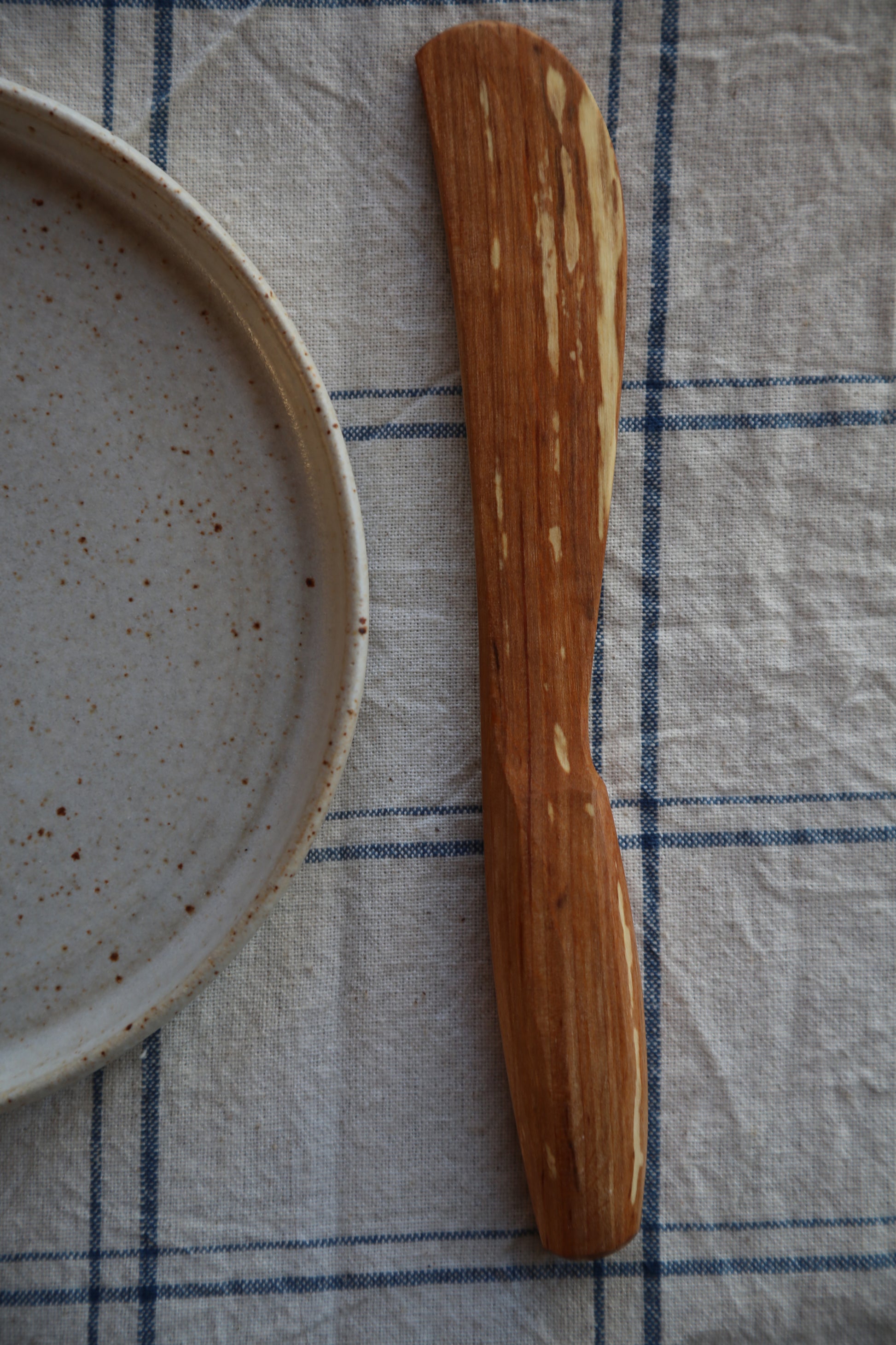 Wooden Butter spreader  on a checkered cloth with a ceramic plate in the background