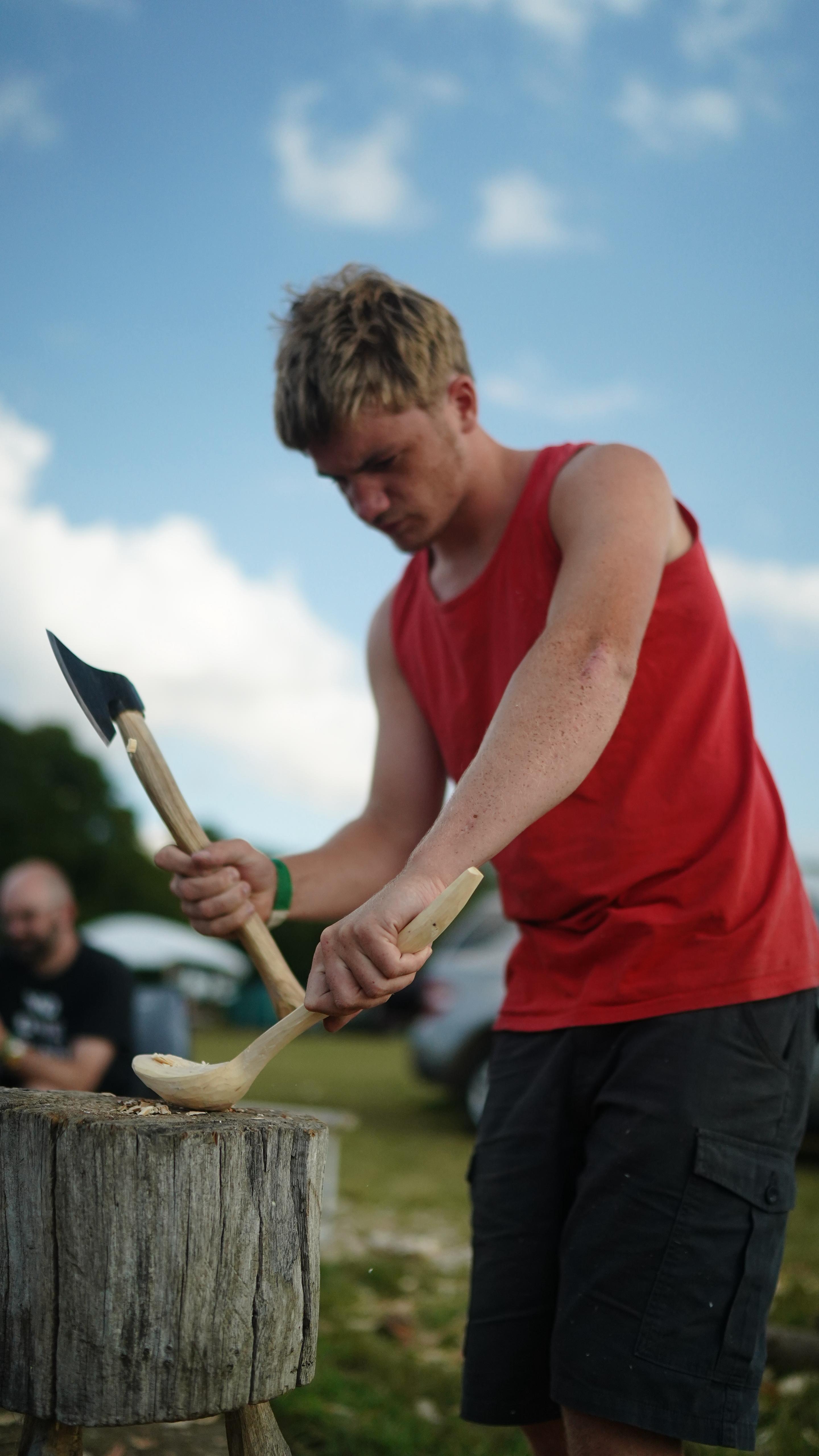 Person in a red tank top using an axe to carve a wooden spoon on a log outdoors with a blue sky.