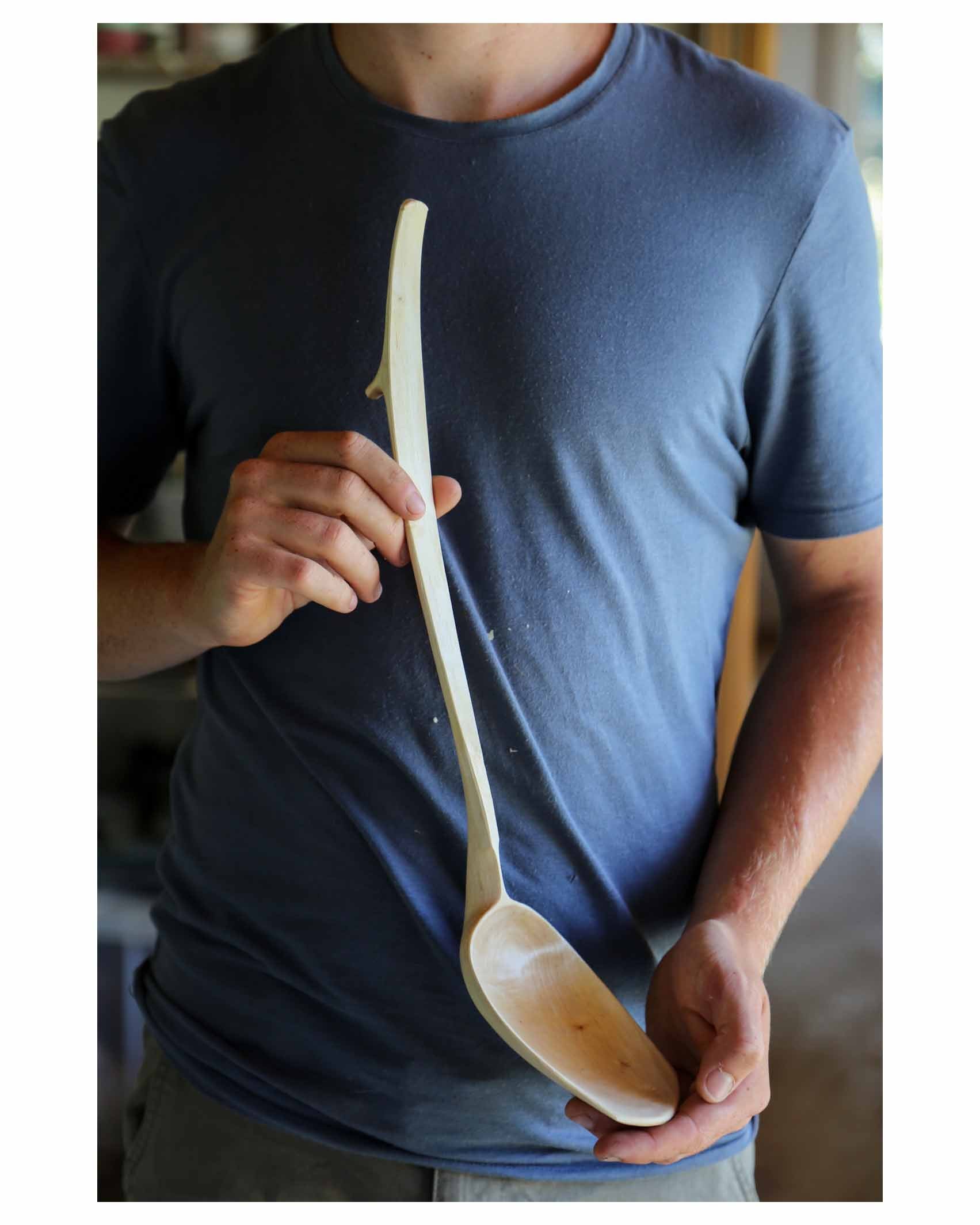 Bent branch birch ladle. handcarved from fallen wood. pale white birch agaisnt a dark blue tshirt held by the hands of the maker