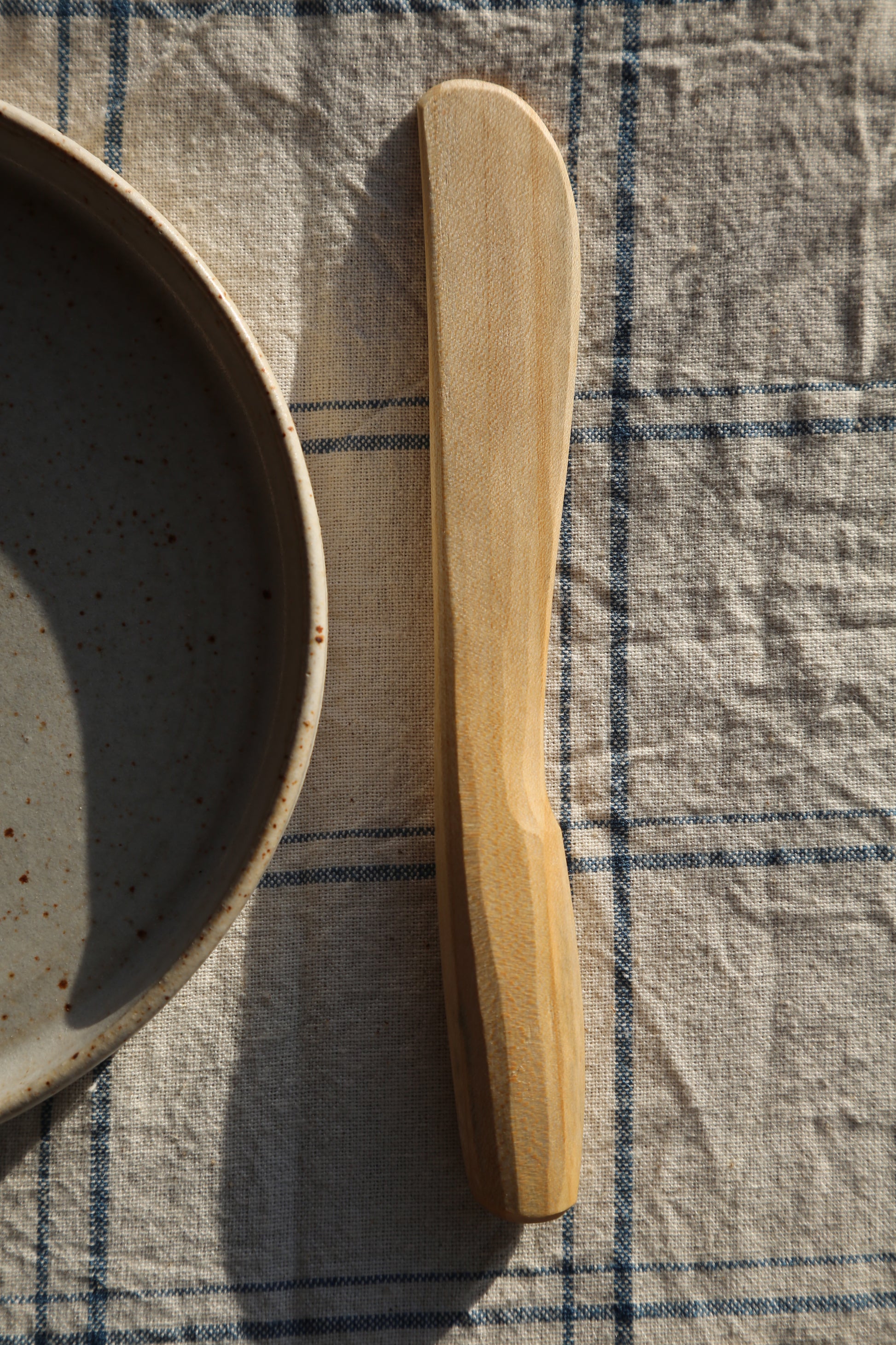Front view of a handcarved sycamore butter spreader with the sun shining down and resting on a clean white stonewear plate 