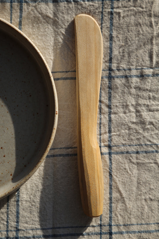 Front view of a handcarved sycamore butter spreader with the sun shining down and resting on a clean white stonewear plate 
